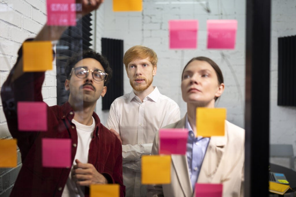 Medium shot people looking at post its. Three peoples are discussing the plan on glass wall.