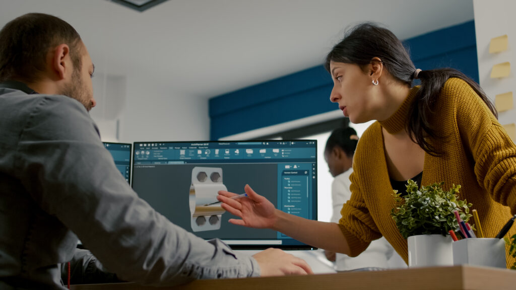 Industrial designer discussing with colleague while working in CAD program, designing 3D prototype of components. Woman engineer using pc with two monitors sharing ideas for construction mechanism